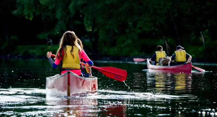 Two people each paddle two red canoes away from the camera. They are wearing yellow life jackets. 